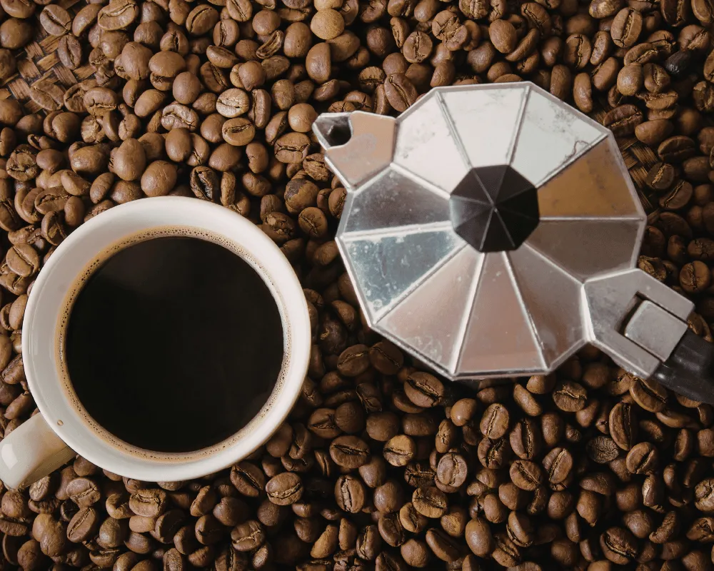Taza de café negro junto a una cafetera italiana moka sobre granos de café tostado.