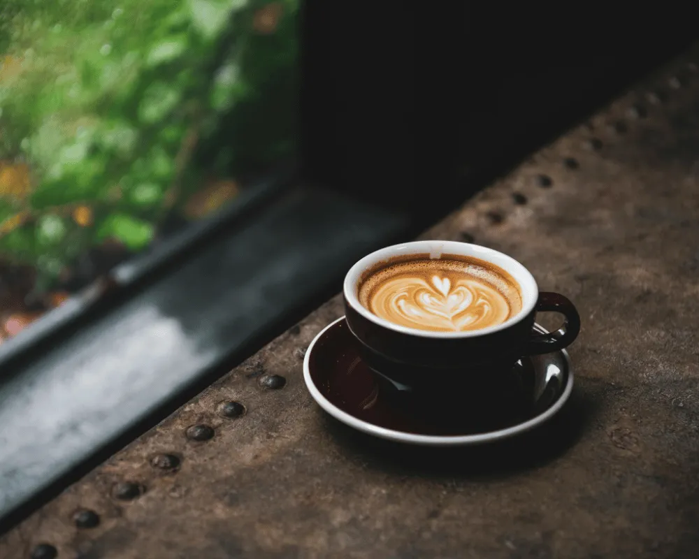 Taza de café arábica con latte art en espuma de leche sobre una mesa de madera junto a una ventana, representando café de especialidad.